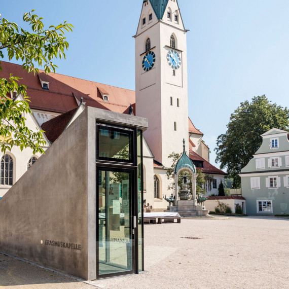 Keilförmiger Eingangsbereich aus Beton zur unterirdischen Erasmuskapelle, dahinter St. Mang Kirche