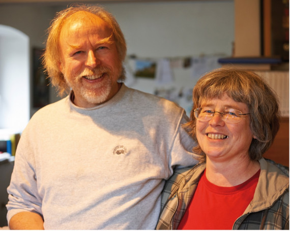 ein älterer lächelnder Herr und eine lachende Frau mit Brille blicken in die Kamera