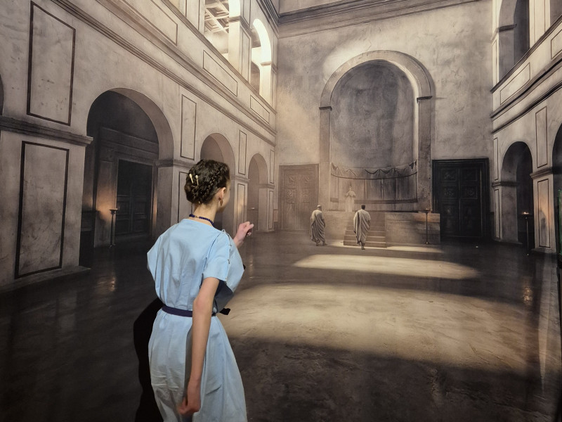 Ein als R&ouml;merin gekleidetes M&auml;dchen betritt scheinbar ein r&ouml;misches Geb&auml;ude, das aber nur ein Druck auf einem deckenhohen Banner ist.